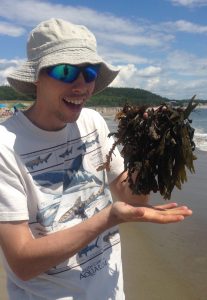 young man on ocean shore holding up a bunch of seaweed