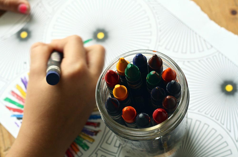 a young child's hand using crayons from a glass jar to color on a coloring sheet