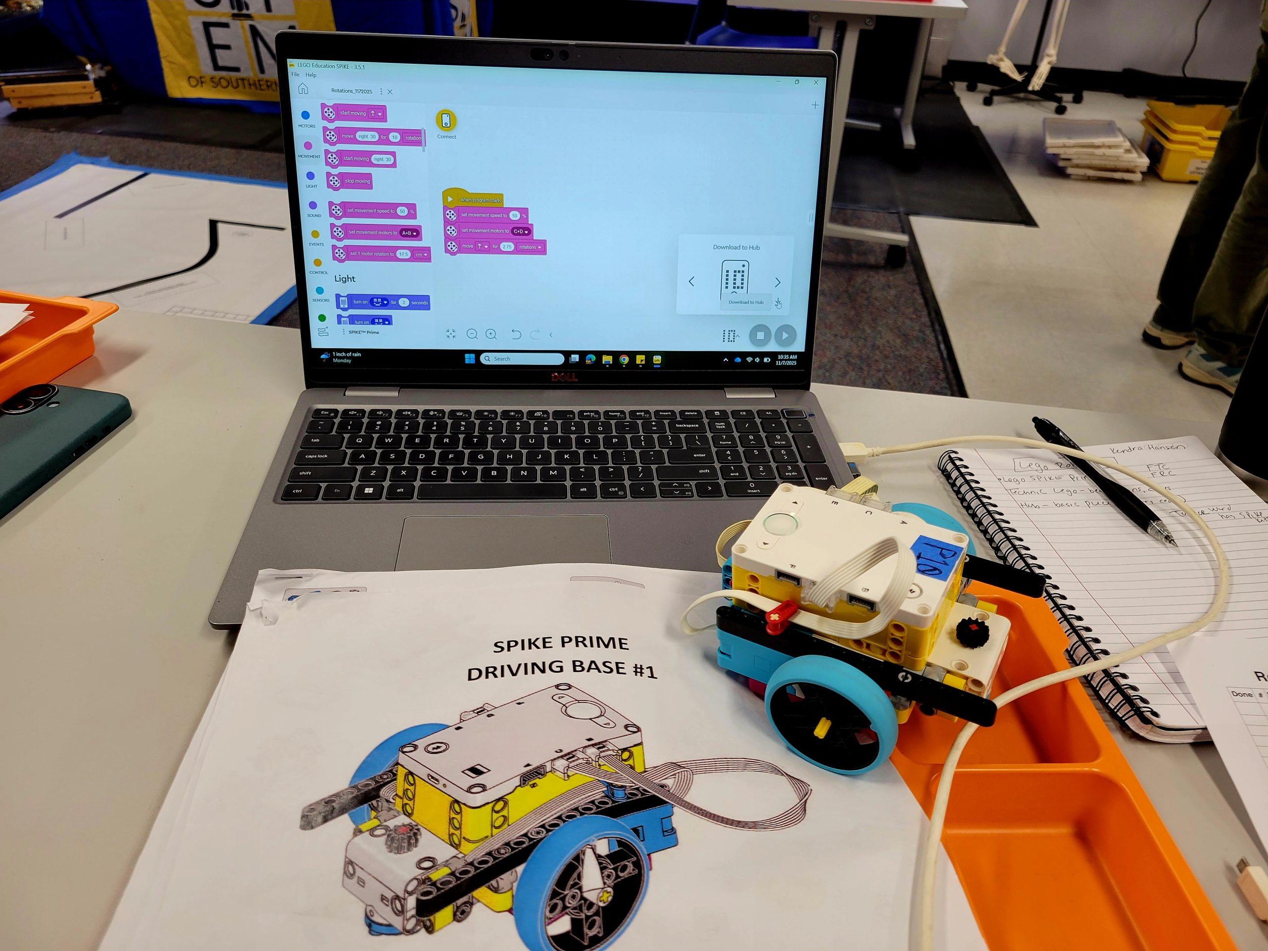 A computer screen open to coding, and a notebook with writing in it, a handbook with spike prime driving base #1 written on it, and a small LEGO driving craft with two large visible wheels.