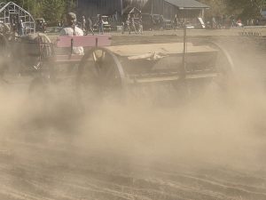 Team of horses pulling historic farming equipment as dust rises from a freshly tilled field during an agricultural demonstration.