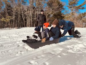 Several kids in one sled smiling and laughing as they are pushed down a snowy hill.