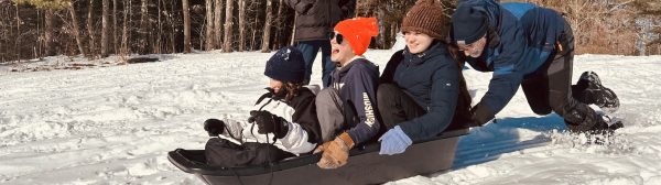 Several kids in one sled smiling and laughing as they are pushed down a snowy hill.