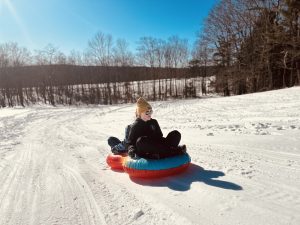 A teen sliding down a hill backwards in a snow tube, wearing sunglasses with a neutral expression.