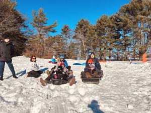 A group of children and adults sitting in the snow and in sleds.