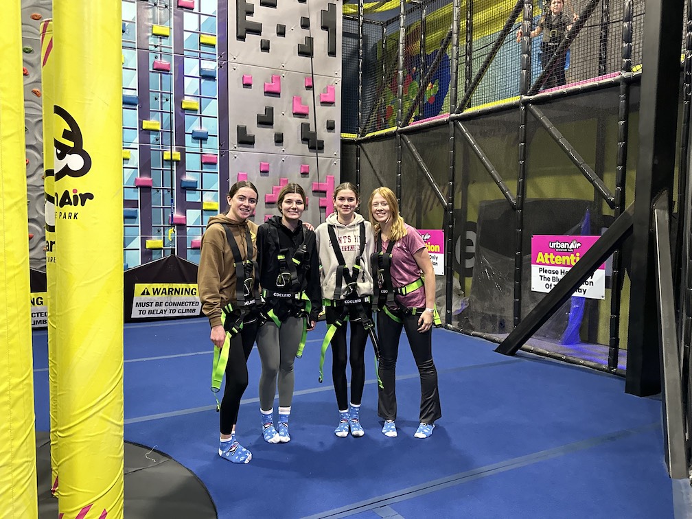 Four GLEE members posing in front of a rock climbing wall, wearing harnesses.