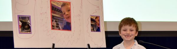 A child smiling in front of a presentation poster about his pet rabbit.