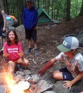 A camper cooking over a open fire while another camper warms her hands nearby.