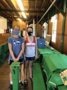 two young girls in facemasks stand next to green table inside hall