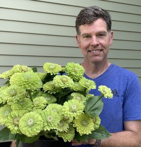 Jason Aucoin, a white man with dark hair, is shown wearing a blue t-shirt and holding a bouquet of chartreuse zinnias from his garden.