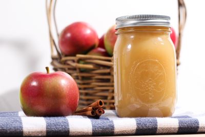 An apple and a jar of homemade applesauce in front of a basket of fresh apples.