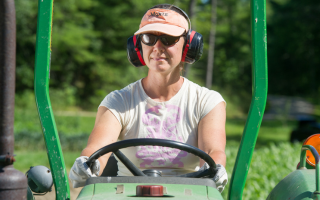 person driving green tractor wearing pink t-shirt, orange hat with the word Moxie printed on it, ear protection, and sunglasses