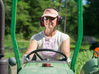 person driving green tractor wearing pink t-shirt, orange hat with the word Moxie printed on it, ear protection, and sunglasses