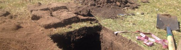 Bleaker Test Pit: sod and soil piled next to a square hole. A shovel and tape measure lay nearby.