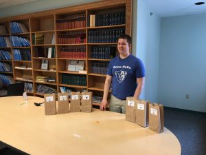 Tyler stands behind a table with 7 small paper bags labeled Site A through G