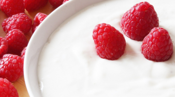 White bowl of yogurt with 3 red raspberries on the surface. Other raspberries visible in the background.