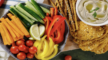 Overhead view of strips of chopped vegetables on a board with a small dish of hummus and crackers