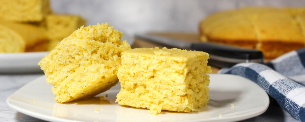 Two corn bread squares on a plate with corn bread loaf in the background.