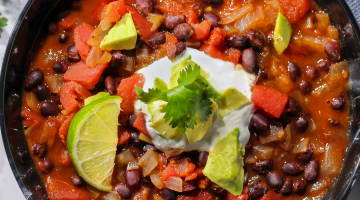 Bowl of black bean soup topped with avocado, sour cream, cilantro, and lime wedges.