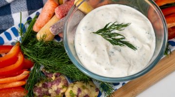 herb yogurt dip in a glass bowl with fresh dill garnish. Placed on a serving tray surrounded by raw red pepper strips, multicolored carrots, and crackers.