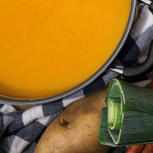 Carrot leek soup in a soup pot with a whole potato, leek and carrots in the background.