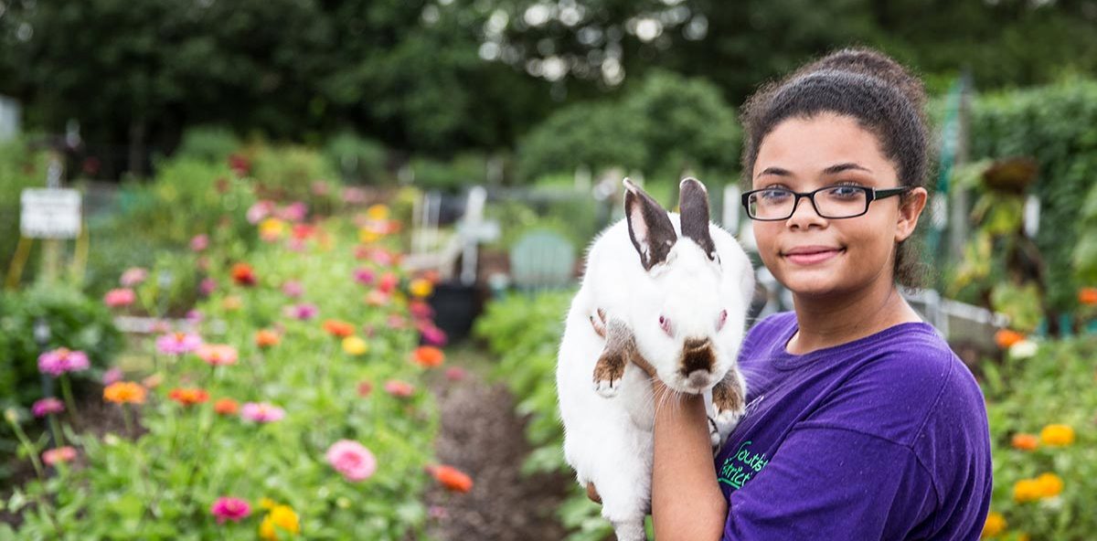 a girl with a rabbit standing in front of a garden