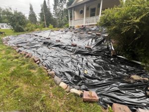 terraced garden area along the edge of a house with a deck is covered with black plastic anchored down with rocks and bricks.