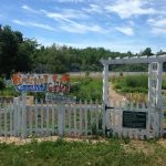 Riverton Community Garden Sign atop white picket fence surrounding a large in ground garden area with paths covered in wood chips.