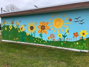 A colorful mural painted on a light blue cinder block building. The mural depicts a garden scene with a variety of colorful flowers, including sunflowers, on a green hill. A large yellow sun with rays shines in the sky. In the upper right corner, a few birds are flying. The bottom of the wall is painted white, and there is a grassy lawn in the foreground. The sky is cloudy and grey.