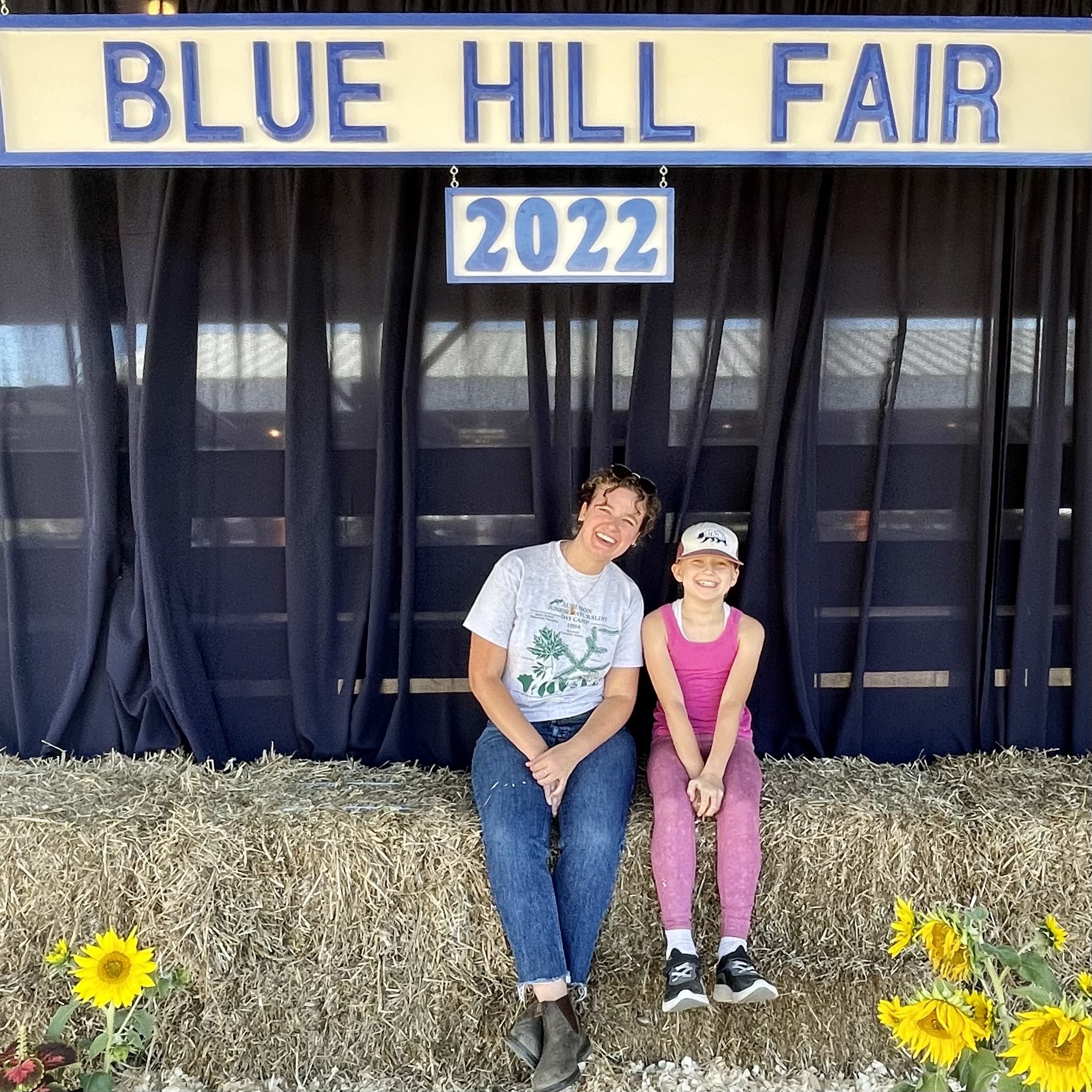 4-H Worker and Student sitting on hay bales under blue hill fair signage