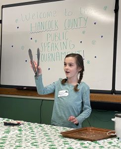 Young girl holding up kitchen tongs during her public speaking demonstration