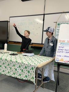 Two 4-H youth presenting how to make a grilled cheese, with one of the youth pointing to the crowd as if she were selecting someone to ask a question