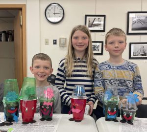 Three young kids (2 boys and 1 girl) standing with their hand made plant pets (plants in cups decorated with faces, hands, feet, bows, etc)