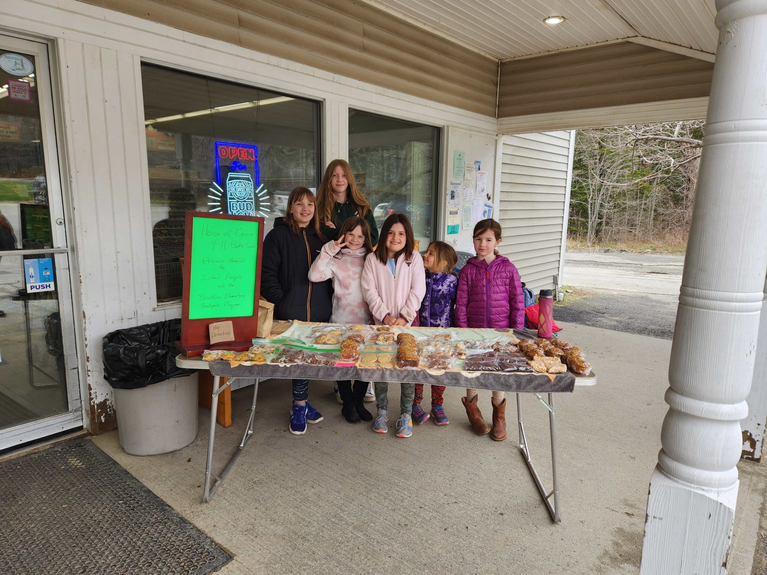 6 girls outside a convenience store with baked goods on a table in front of them.