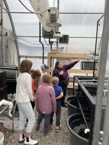 5 kids grouped around an adult pointing out aquaponics plumbing in a greenhouse