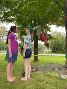 Two girls comparing height in the foreground and one girl hanging upside down from a slackline between two trees in the background
