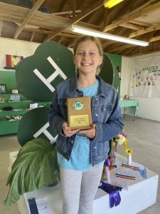 Girl smiling at the camera standing in front of a large 4-H clover and holding up her award for gardening