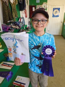 Young boy holding up his handmade Dogman comic and best project ribbon, smiling widely at camera