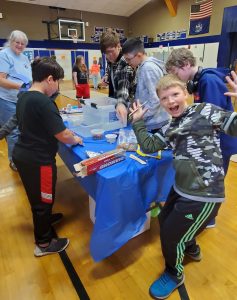 Group of youth gathered around a table using miscellaneous materials to build boats. One youth is smiling animatedly at the camera with his hands up.