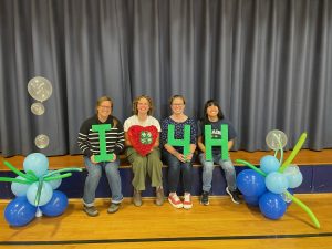 4-H Staff sitting on stage holding "I - Heart - 4 - H" signs