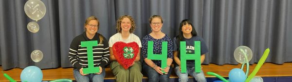 4-H Staff sitting on stage holding "I - Heart - 4 - H" signs