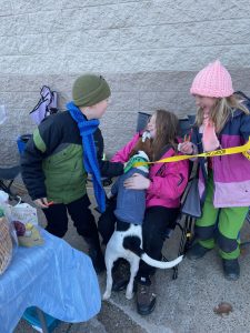 Three youth gathered around a puppy. The puppy is jumping up on the middle child and all are smiling.