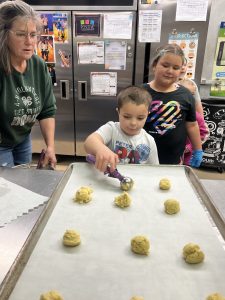 One youth using a scoop to put cookie balls on a tray. Another youth looks on form behind and an adult advises from the sidelines.