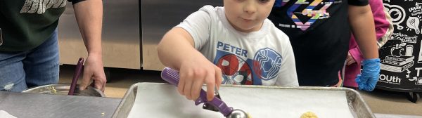 One youth using a scoop to put cookie balls on a tray. Another youth looks on form behind and an adult advises from the sidelines.