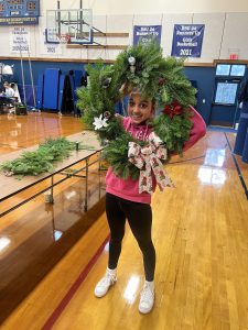 Youth holding up a big green wreath with a bow decoration. She smiles through the middle of it.