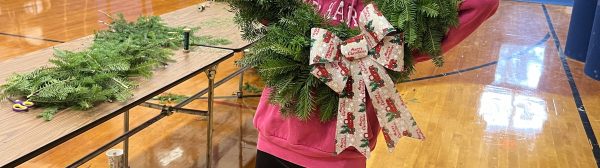 Youth holding up a big green wreath with a bow decoration. She smiles through the middle of it.