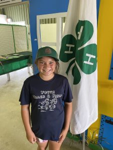 4-H youth wearing a hat standing in front of a 4-H clover flag smiling