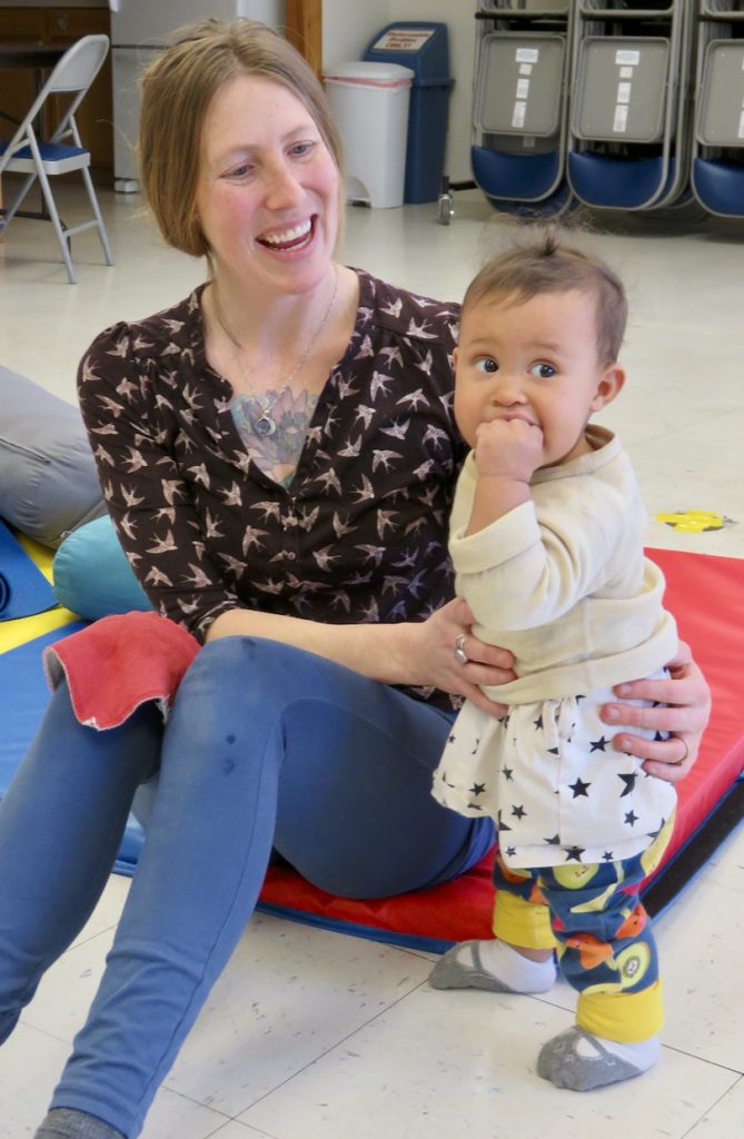 woman in workout clothes sitting on an exercise mat supporting a toddler standing next to her