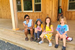 three children and one adult sitting on the porch steps of a wooden building, another child standing behind them in a doorway