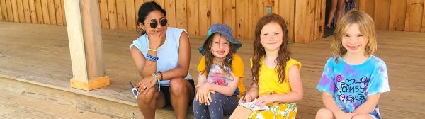 three children and one adult sitting on the porch steps of a wooden building, another child standing behind them in a doorway