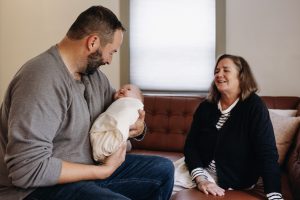 Man sitting on a couch holding a baby doll wrapped in a blanket, woman sitting nearby watching